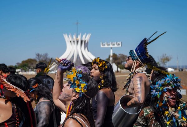 Mulheres indígenas Marcham em Brasília. no primeiro plano, está, em destaque, uma mulher com olhos fechados e cantando. Ao fundo, depois de outras mulheres, se vê a Catedral de Brasília. 