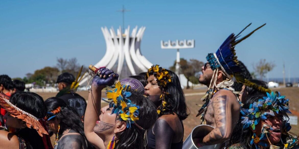Mulheres indígenas Marcham em Brasília. no primeiro plano, está, em destaque, uma mulher com olhos fechados e cantando. Ao fundo, depois de outras mulheres, se vê a Catedral de Brasília. 